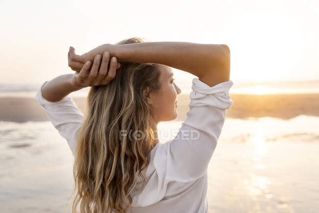Mujer despreocupada con las manos detrás de la cabeza mirando a la vista mientras está de pie contra el mar - foto de stock