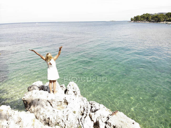 Жінка з піднятими руками дивиться на море, стоячи на скелі в сонячний день. — стокове фото