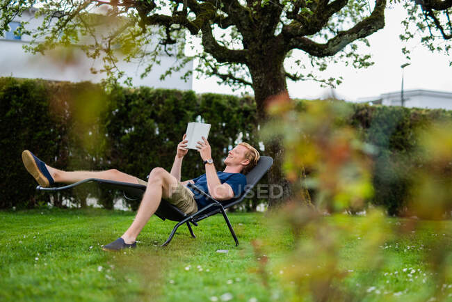 Щасливий чоловік читає книжку, лежачи на стільці на задньому подвір 