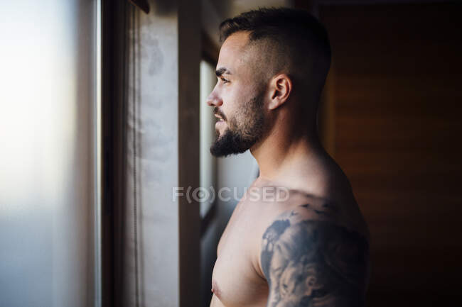 Hombre sin camisa con tatuaje mirando a través de la ventana en casa - foto de stock