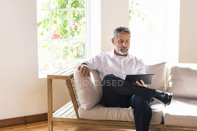 Homme d'affaires mature utilisant une tablette numérique assis avec les jambes croisées au genou sur le canapé à la maison — Photo de stock