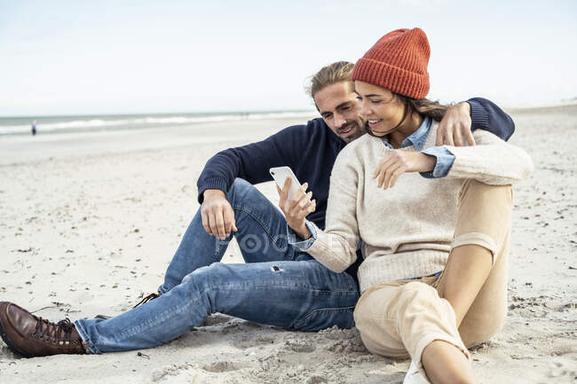 Jeune couple assis ensemble sur le sable de la plage et en utilisant un téléphone intelligent — Photo de stock