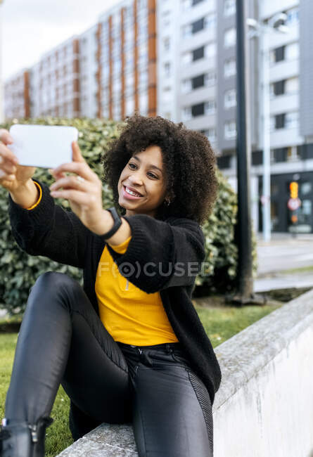Sonriente hermosa mujer tomando selfie con teléfono móvil mientras está sentado en barandilla en la ciudad - foto de stock