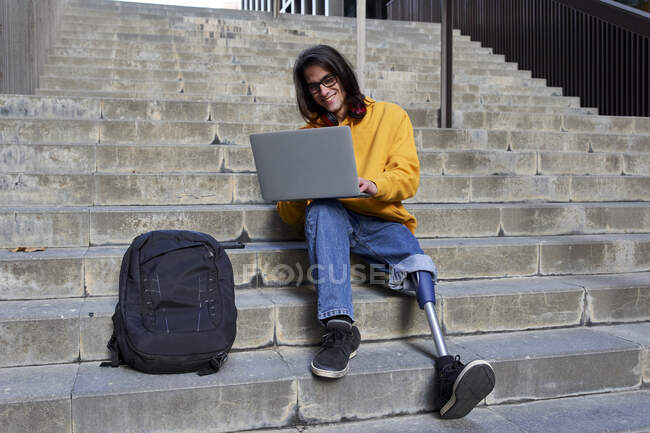 Homem sorridente com prótese de perna usando laptop enquanto sentado em passos — Fotografia de Stock