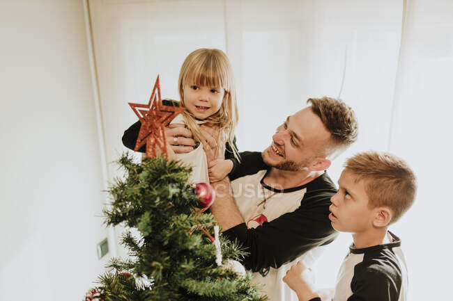 Father showing Christmas tree decoration while carrying daughter by son against wall at home — Stock Photo