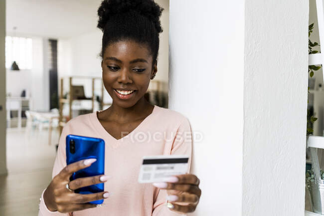 Woman with credit card smiling while using mobile phone at home — Stock Photo
