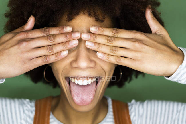 Alegre joven mujer sobresaliendo lengua mientras cubre los ojos contra la pared verde - foto de stock