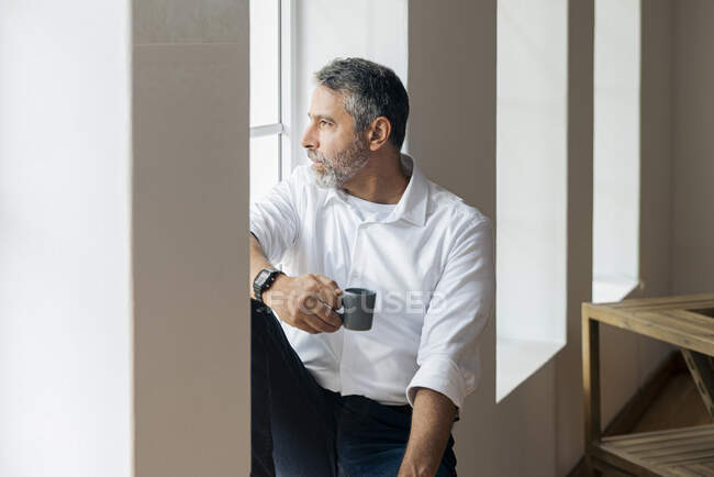 Homme d'affaires réfléchi avec tasse de café regardant par la fenêtre tout en étant assis à la maison — Photo de stock