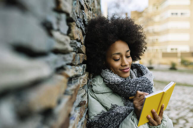 Улыбающаяся афро-женщина пишет в книге, опираясь на каменную стену зимой — стоковое фото