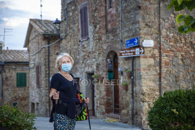 Mujer mayor con mascarilla protectora sosteniendo bastón de senderismo mientras está de pie en la ciudad italiana - foto de stock