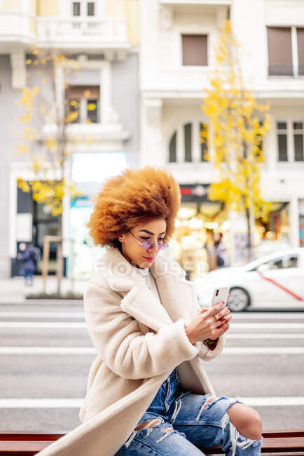 Mujer con cabello rubio afro usando un teléfono inteligente mientras está sentada en el banco de la ciudad - foto de stock