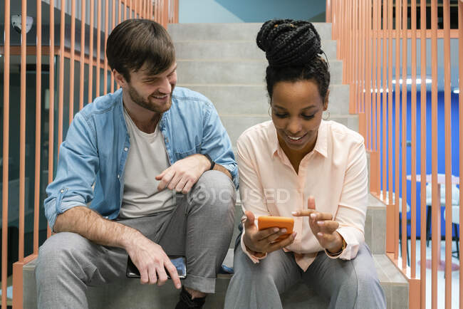Des hommes d'affaires souriants utilisant un téléphone portable et parlant assis sur un escalier au travail — Photo de stock