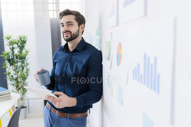 Imprenditore maschio sorridente con tablet digitale che prende il caffè mentre si appoggia alla parete in ufficio — Foto stock