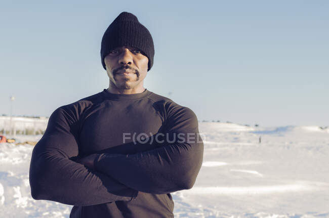 Costruire muscolare sportivo indossando cappello maglia in piedi con le braccia incrociate nella neve durante la giornata di sole — Foto stock