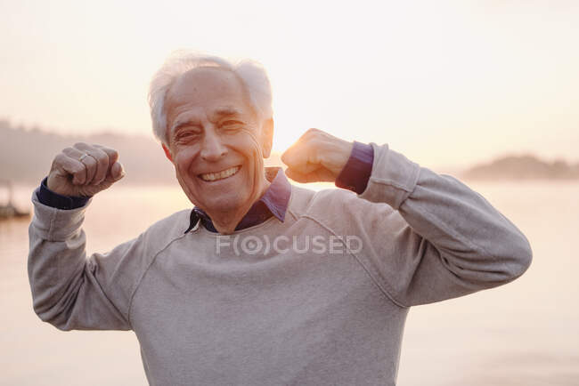 Hombre mayor sonriente mostrando bíceps mientras está de pie contra el cielo durante el amanecer - foto de stock