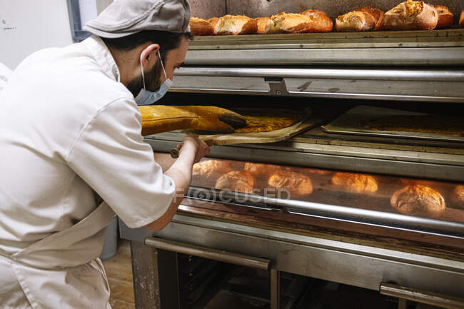Bäcker mit Pizzaschalen überprüft Brot im Backofen während COVID-19 — Stockfoto