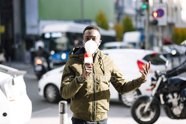 Maschio reporter TV indossando maschera facciale parlando sopra il microfono mentre in piedi sulla strada — Foto stock