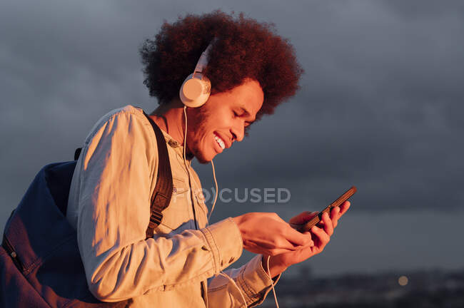 Uomo sorridente ascoltare musica e utilizzando smartphone contro cielo nuvoloso al tramonto — Foto stock