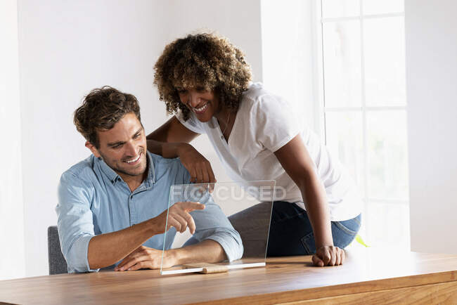 Усміхнена пара торкається прозорого екрану пристрою, сидячи вдома. — стокове фото