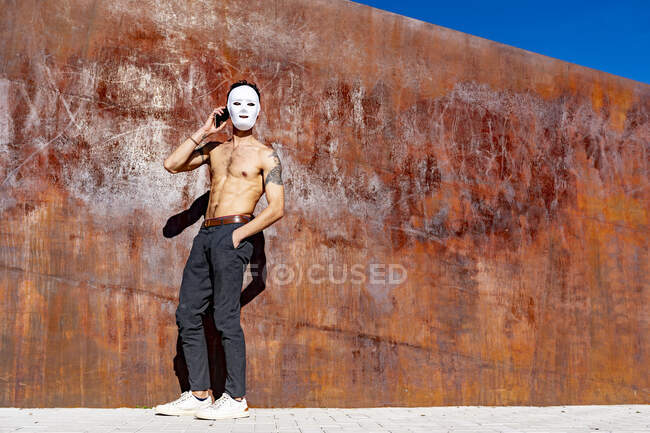 Hombre con máscara blanca hablando en el teléfono móvil mientras está de pie con las manos en los bolsillos contra la pared - foto de stock