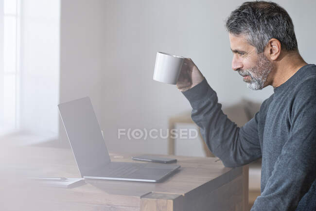 Empresario con taza de café hablando en videollamada a través de computadora portátil mientras está sentado en casa - foto de stock