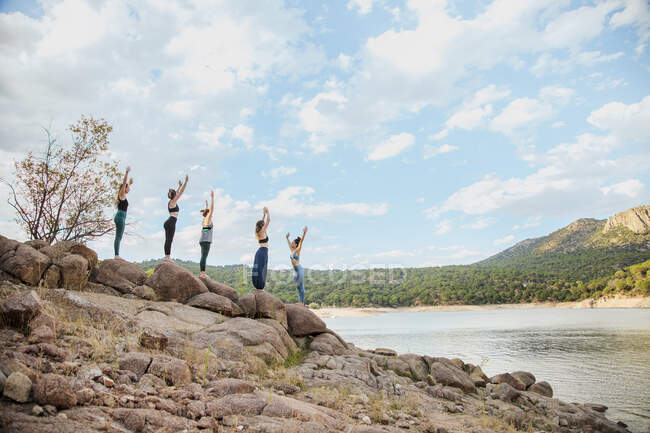 Mujeres jóvenes practicando yoga con instructor sobre roca contra cielo - foto de stock