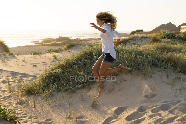 Mujer despreocupada saltando sobre la arena en la playa al atardecer - foto de stock