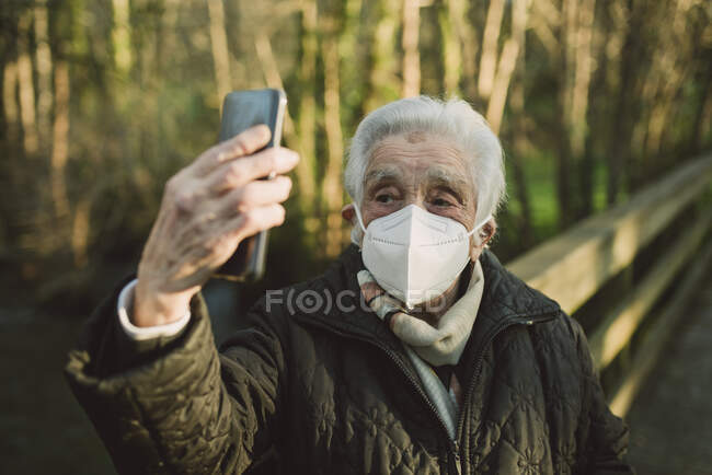 Старшая женщина носит защитную маску для лица, когда делает селфи через смартфон зимой — стоковое фото
