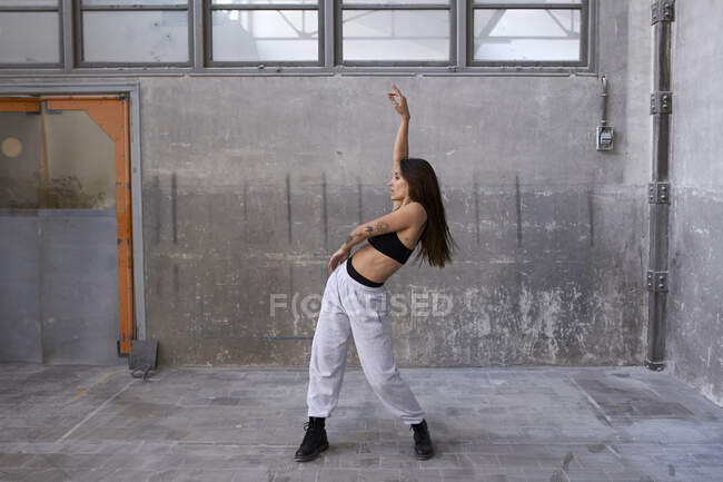 Ballerina che balla con il braccio alzato contro il muro grigio in fabbrica abbandonata — Foto stock