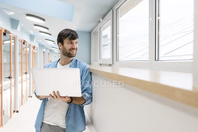 Улыбающийся бизнесмен смотрит в окно, держа ноутбук в офисном коридоре — стоковое фото