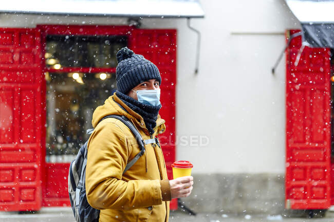 Людина з чашкою кави стоїть проти магазину під час снігопаду під час пандемії — стокове фото