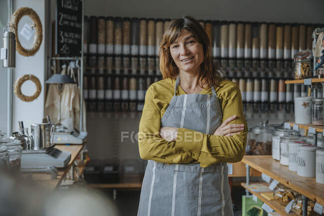 Happy female store clerk with arms crossed standing in retail shop — Stock Photo