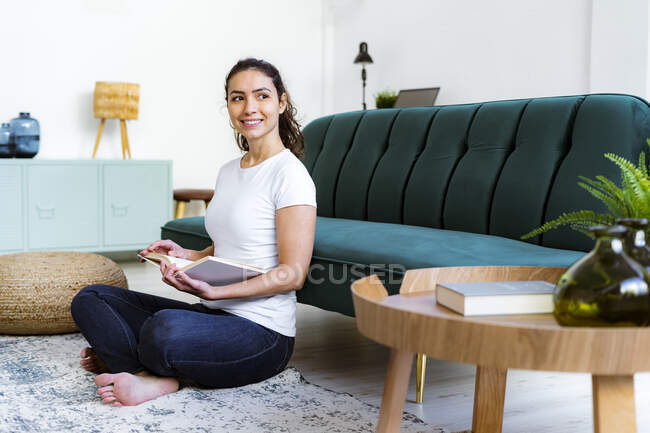 Mujer sonriente con libro mirando hacia otro lado mientras está sentada en casa - foto de stock
