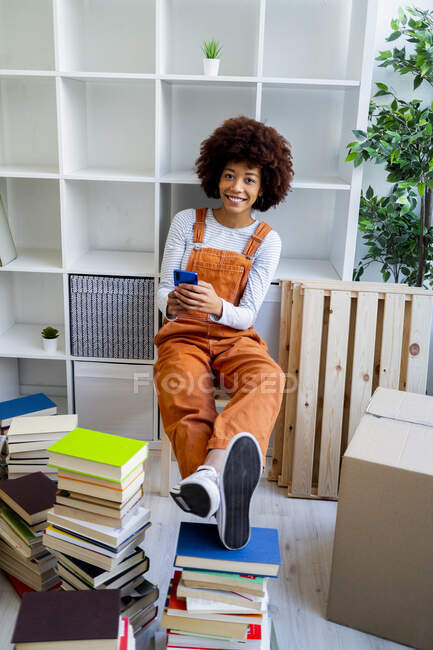 Mujer afro sonriente sosteniendo el teléfono móvil mientras se sienta contra la estantería en un nuevo hogar - foto de stock