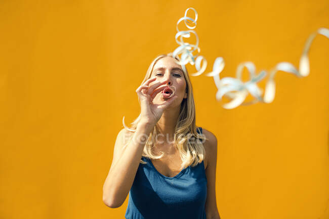 Schöne Frau bläst an sonnigem Tag Konfetti gegen gelbe Wand — Stockfoto
