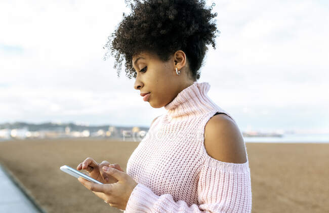 Belle femme avec les cheveux bouclés en utilisant un téléphone mobile tout en étant assis contre le ciel clair — Photo de stock