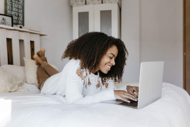 Giovane donna sorridente mentre si utilizza il computer portatile sdraiato sul letto a casa — Foto stock