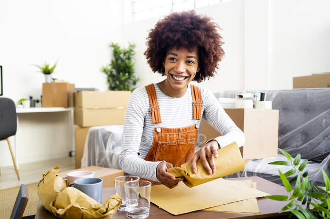 Happy Afro young woman wrapping drinking glass in brown paper at new home — Stock Photo