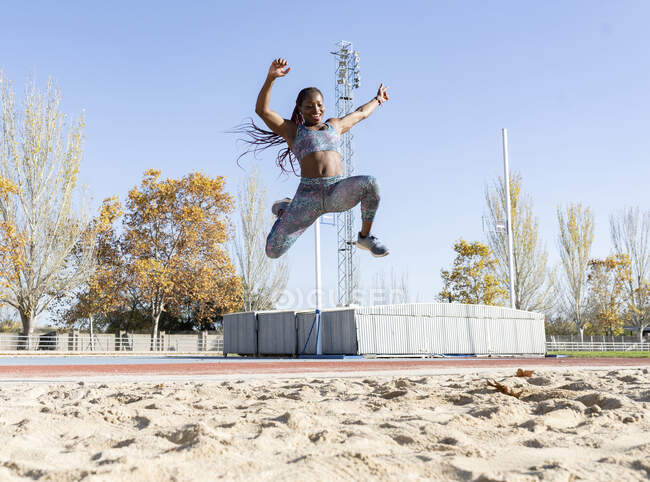 Cheerful sportswoman training for long jump over sand during sunny day — Stock Photo
