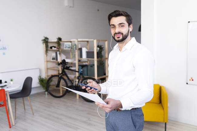 Profesional masculino sonriente con documento en papel que sostiene el teléfono móvil mientras trabaja en el lugar de trabajo - foto de stock