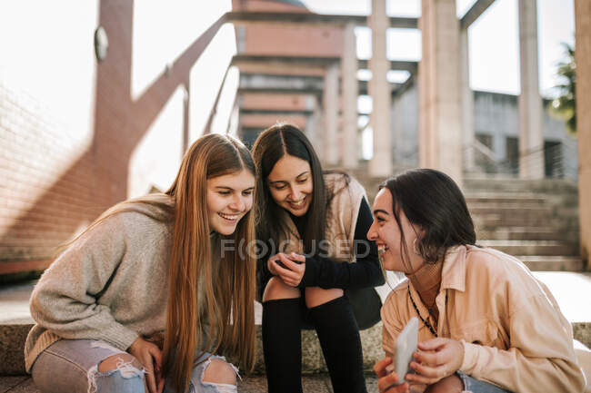 Весела дівчина - підліток, яка показує друзям смартфон, сидячи на сходах у місті. — стокове фото