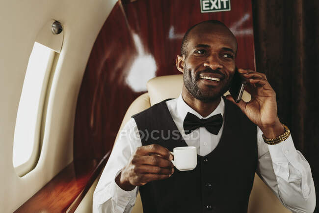Young businessman with coffee cup talking on smart phone in airplane — Stock Photo