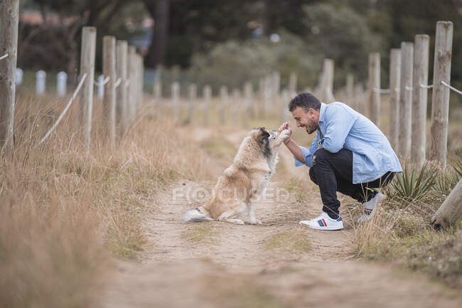 Взрослый мужчина играет с собакой, приседая на грунтовой дороге — стоковое фото