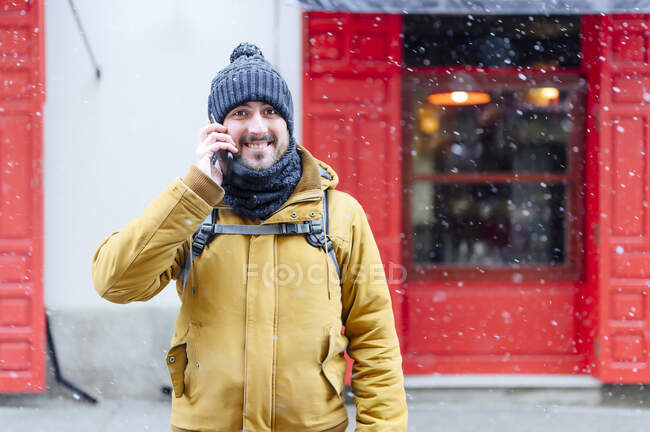 Smiling man in warm clothing talking on smart phone while snowing during winter — Stock Photo