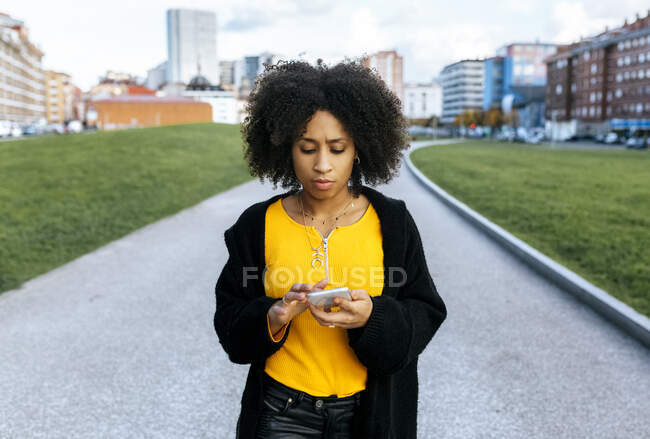 Mujer joven con cabello afro usando teléfono móvil mientras está de pie en el sendero de la ciudad - foto de stock
