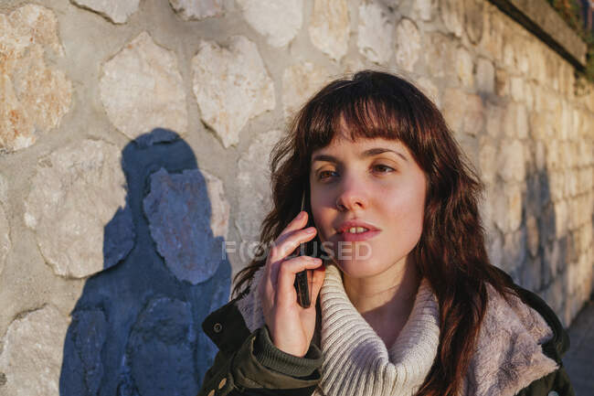 Созерцание молодой женщины, разговаривающей по смартфону о каменной стене во время заката — стоковое фото