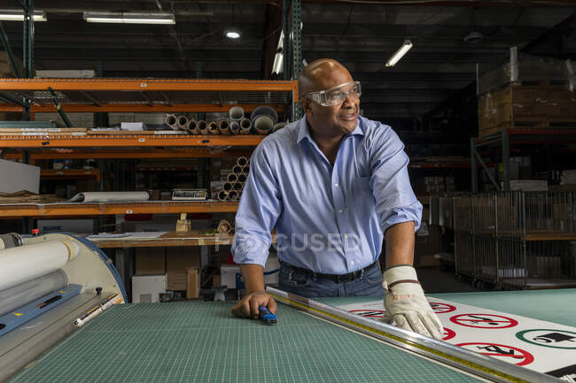 Funcionário sorrindo vestindo óculos de proteção olhando para longe enquanto trabalhava na loja de impressão — Fotografia de Stock