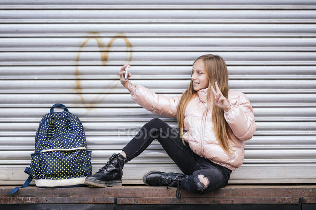 Улыбающаяся девушка в теплой одежде делает селфи через мобильный телефон, жестикулируя против затвора — стоковое фото
