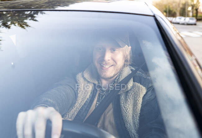 Sonriendo hombre adulto medio conduciendo coche visto a través del parabrisas - foto de stock