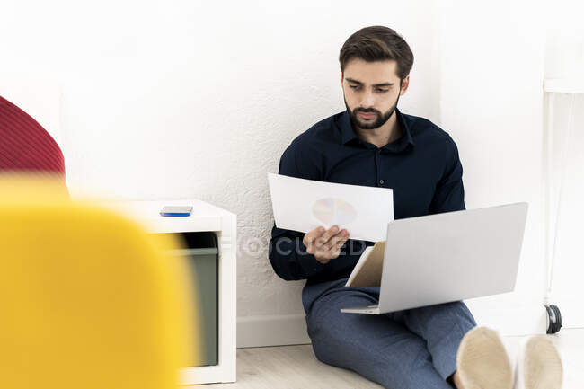 Joven hombre de negocios analizando diagramas con computadora portátil mientras está sentado contra la pared en la oficina - foto de stock
